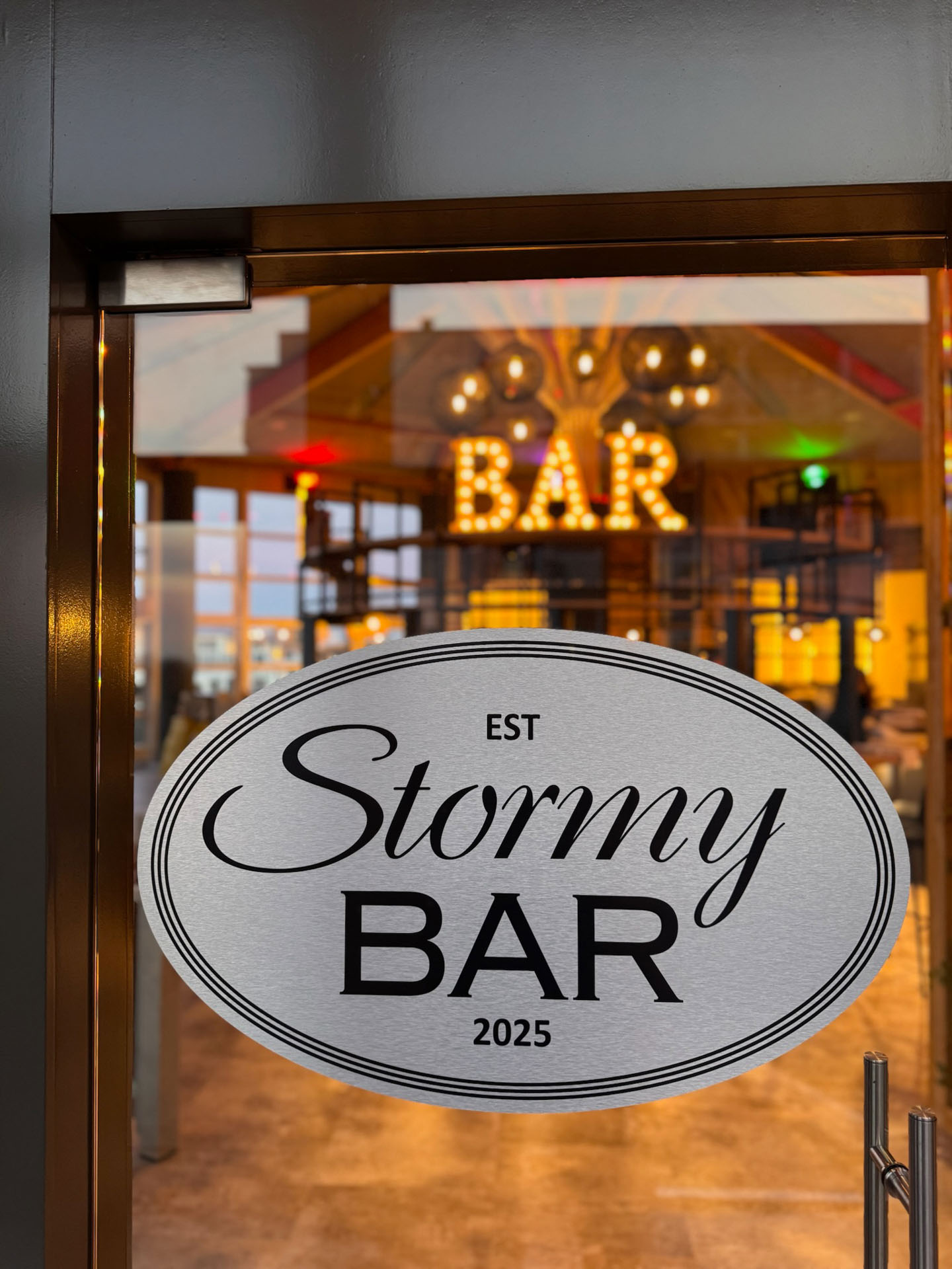 Ovales Schild an Glastür mit „Stormy BAR“ und Innenansicht des beleuchteten Barbereichs.