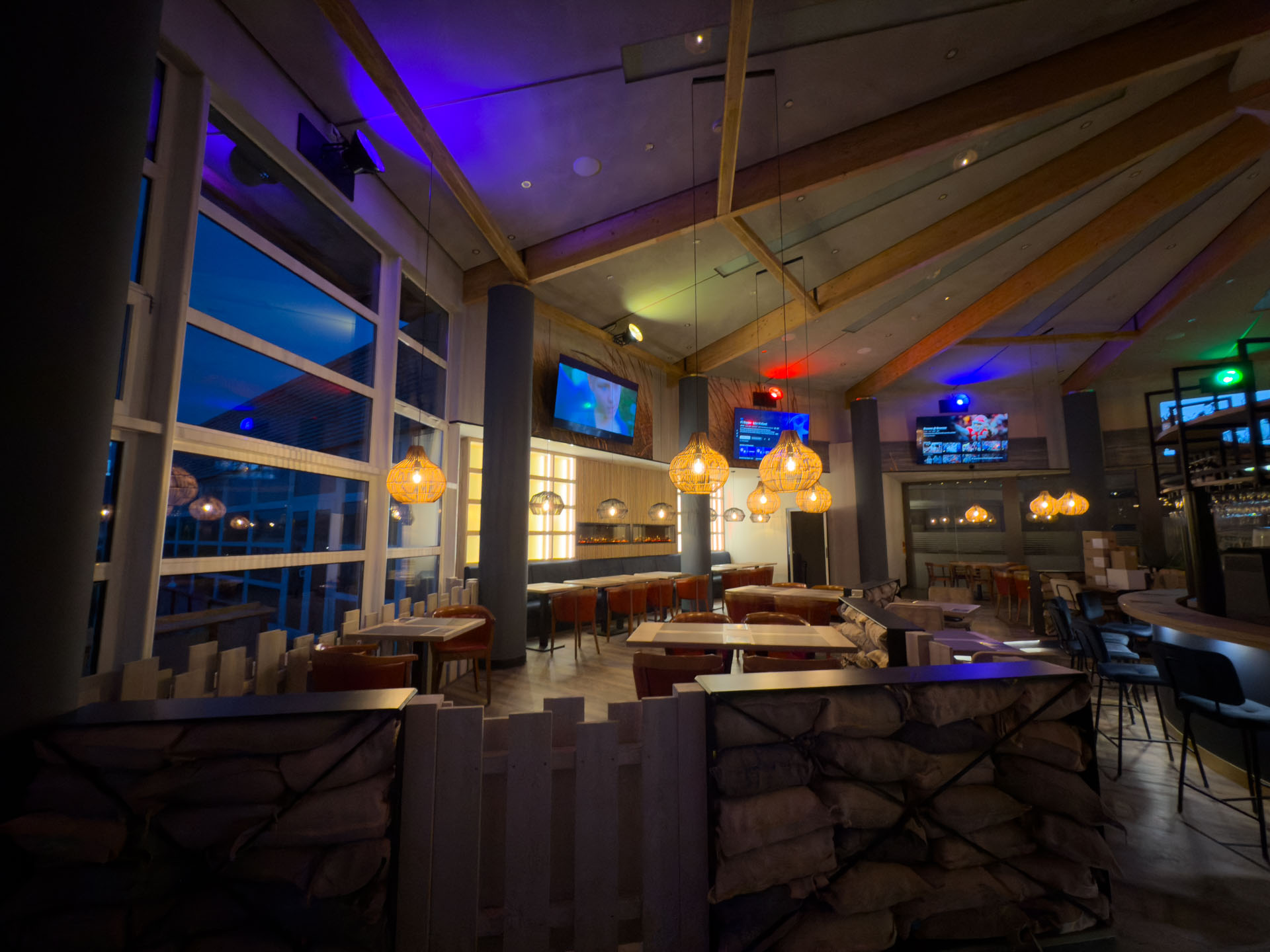 StormyBAR mit Holzbalken, hängenden Lampen und großen Fenstern bei Nachtbeleuchtung.