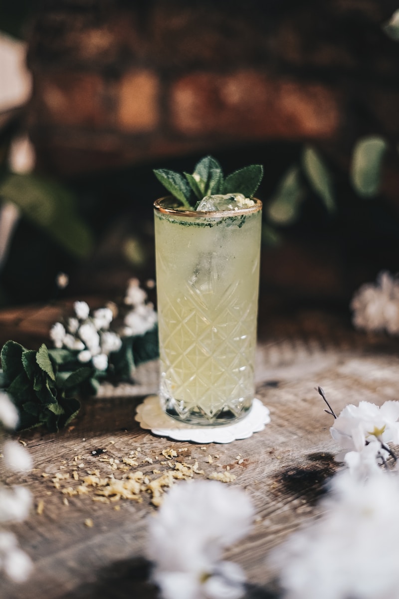 Hoher Cocktail mit Kräutern und Eis, umgeben von weißen Blumen auf Holztisch.