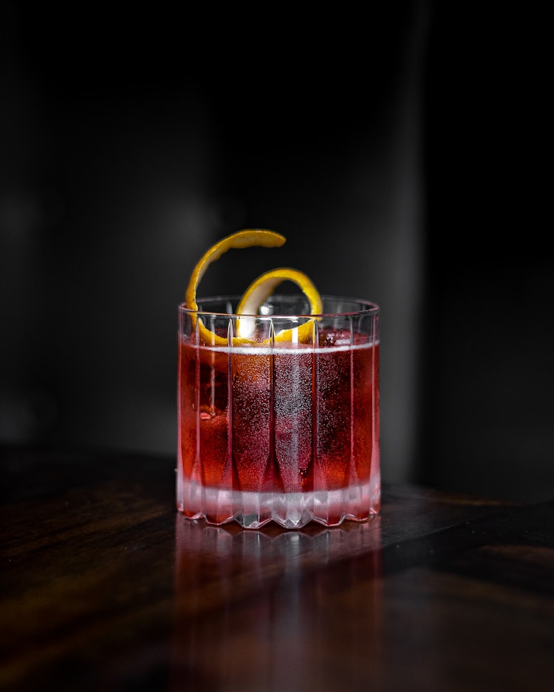 Ein roter Cocktail mit Orangenschale in einem geriffelten Glas auf Holztisch.